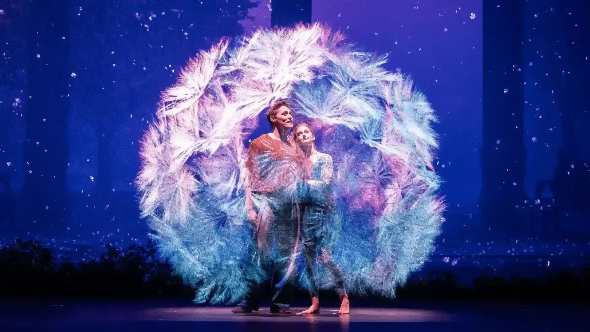 two performers embracing on stage inside a giant projected dandelion, its white and blue seeds drifting against a starry purple backdrop
