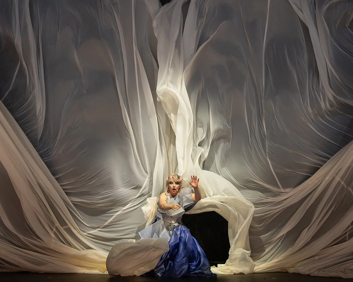a singer in a blue gown kneeling on a dark stage, swallowed by billows of white fabric rising behind her