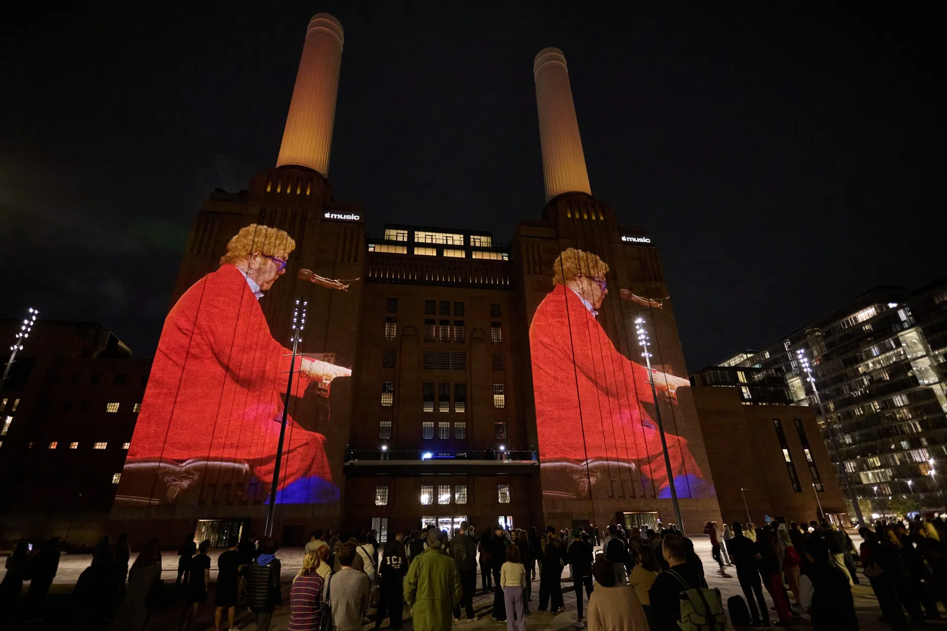 the two towers of Battersea Power Station at night, each carrying a projected image of a pianist in a red suit, a crowd watching below