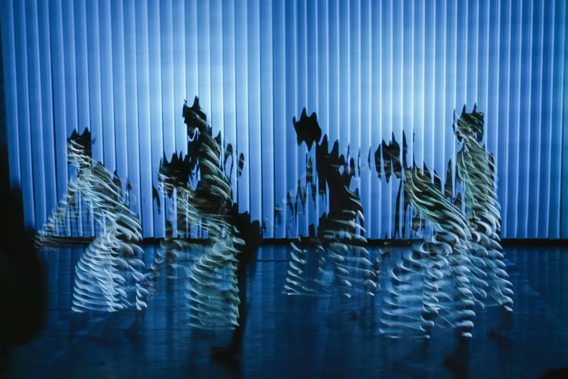 dancers in motion before a pale blue striped backdrop, each body multiplied into a trail of ghostly echoes