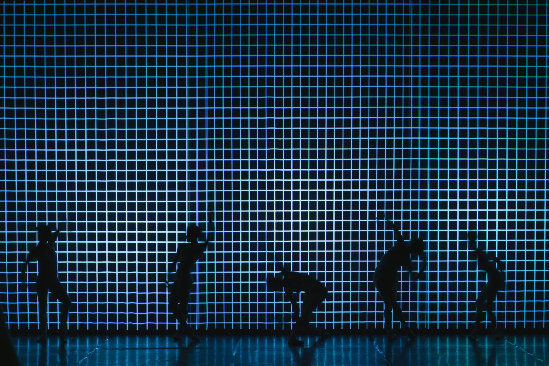 five dancers in silhouette against a wall-high grid of glowing cyan lines