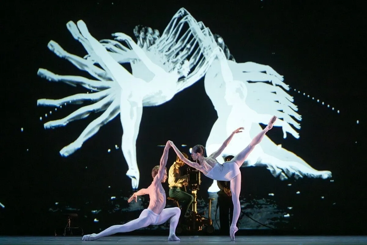 two dancers in white on stage, a camera operator behind them, and on the black screen their silhouettes multiplied into trails of white light