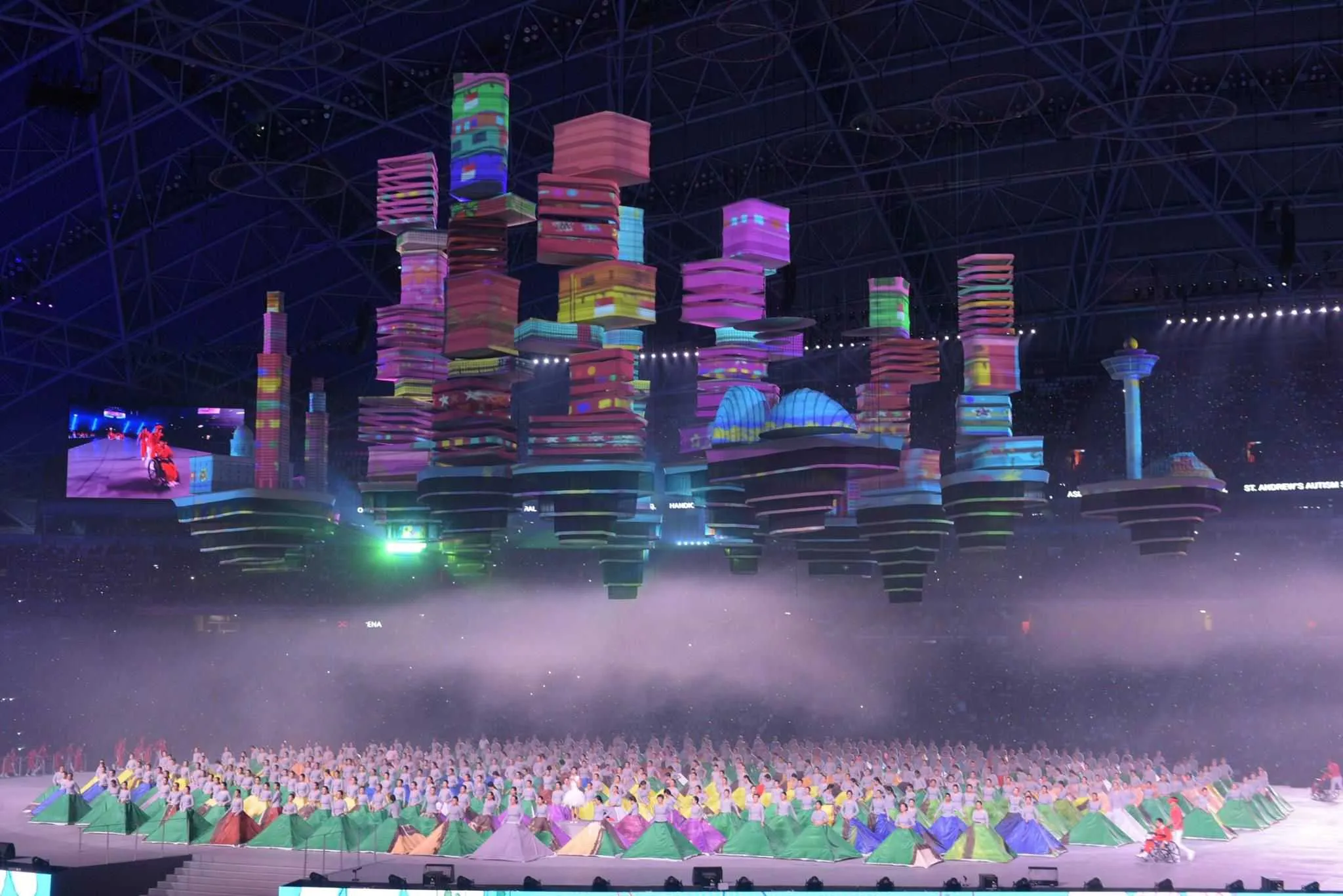the flying city of Singapore National Day: dozens of suspended building blocks, each projection-mapped in vivid colours, above a stadium in haze