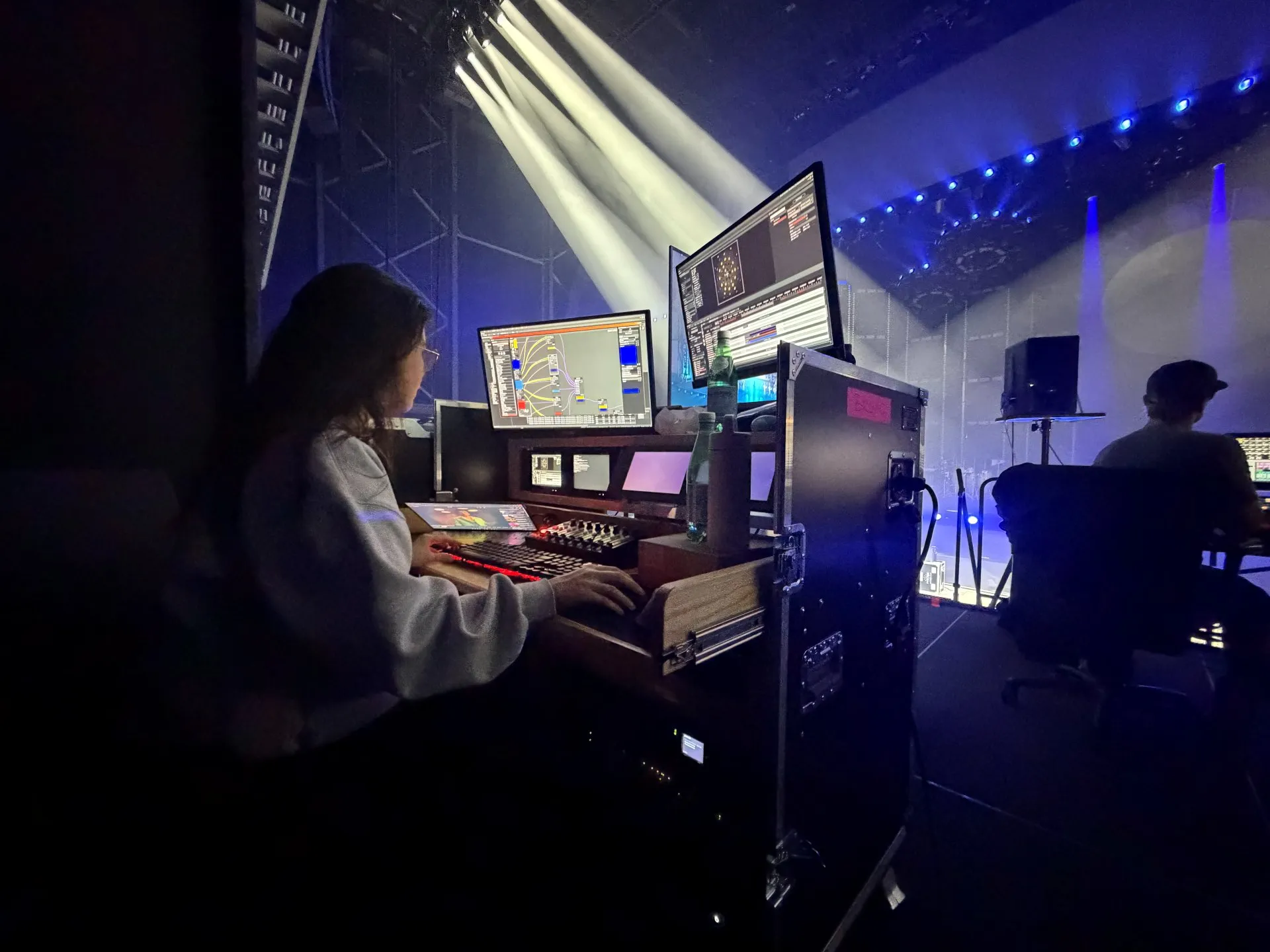 a programmer at the video console in the dark, facing screens and a control surface, light beams crossing the stage behind