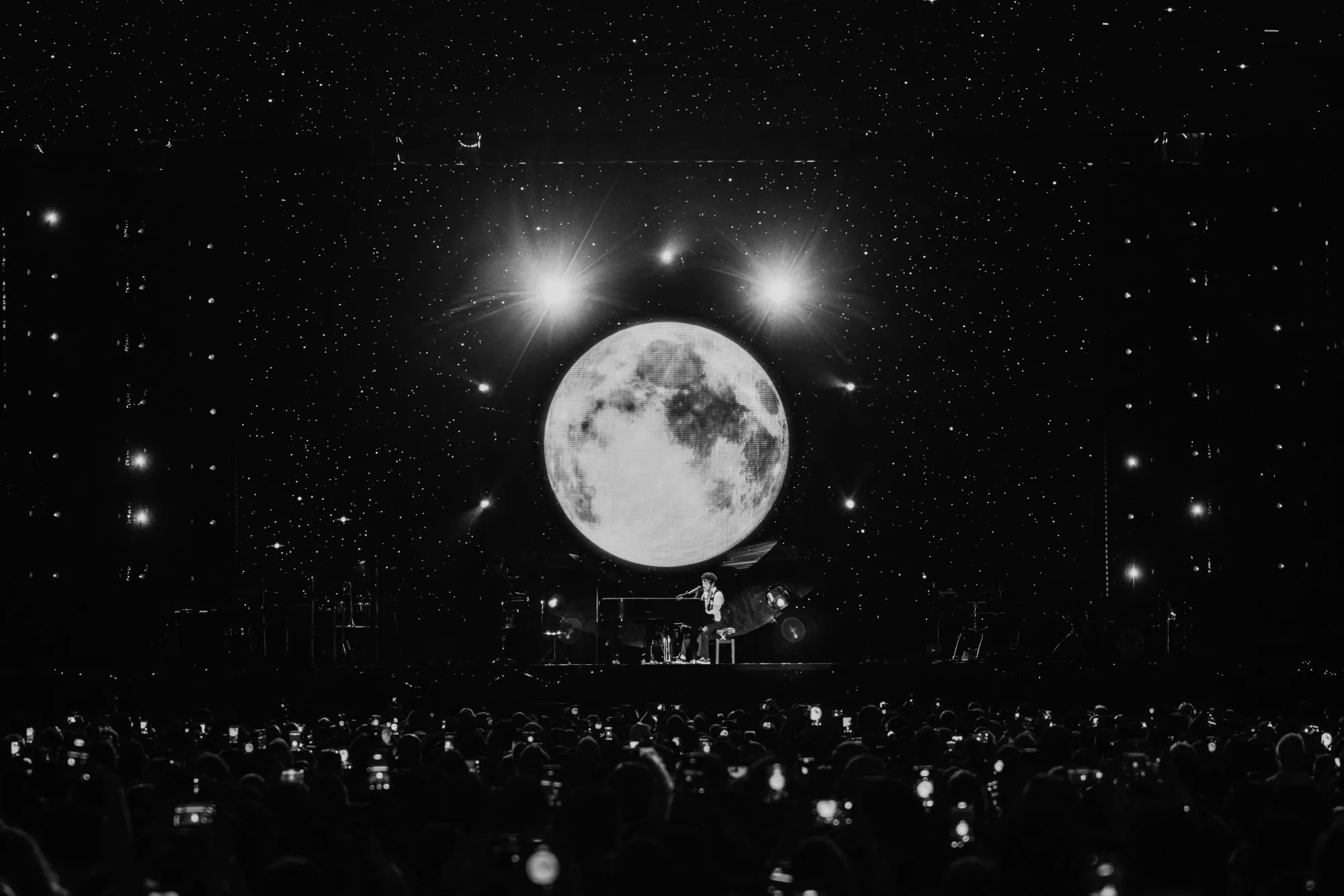 a performer alone at a piano on a dark stage, beneath a full moon filling the screen and a field of stars, the audience below holding up their phones
