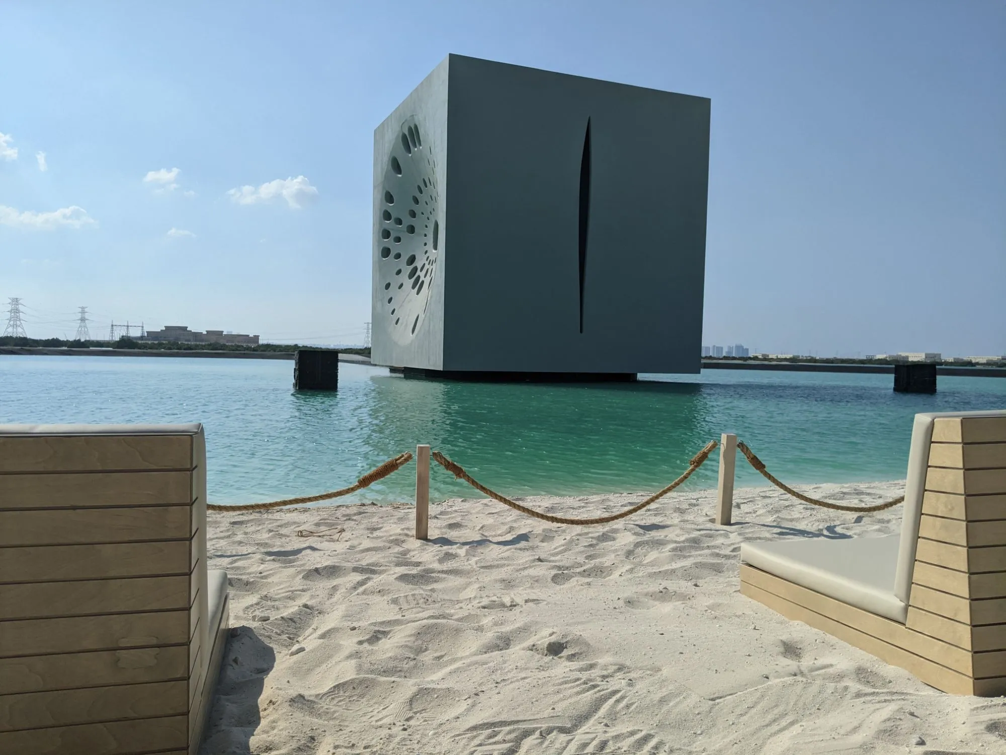 the cube by day standing on turquoise water, seen from the beach between two loungers, a round pattern of openings on one face and a long slit on the other