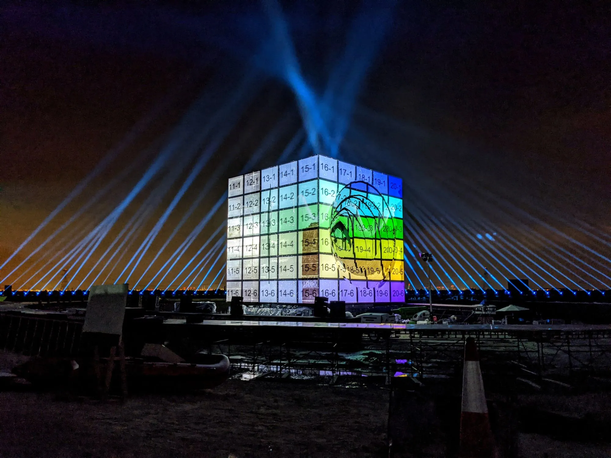 the cube at night covered in a grid of numbered coloured patches, one per projector zone, searchlight beams fanning out behind it