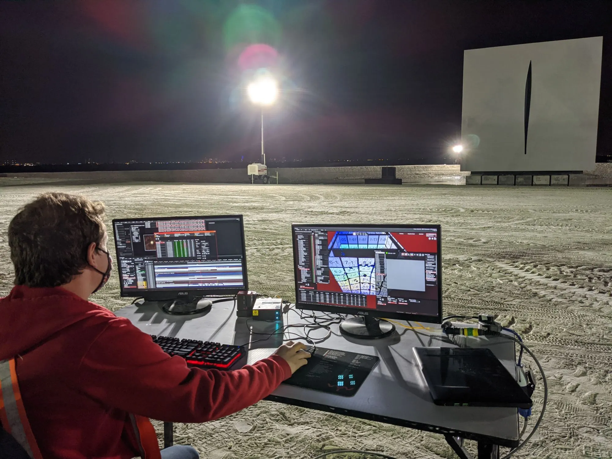 a VYV programmer at two screens on a table on the sand, the PHOTON interface showing the cube, the white cube lit in the distance