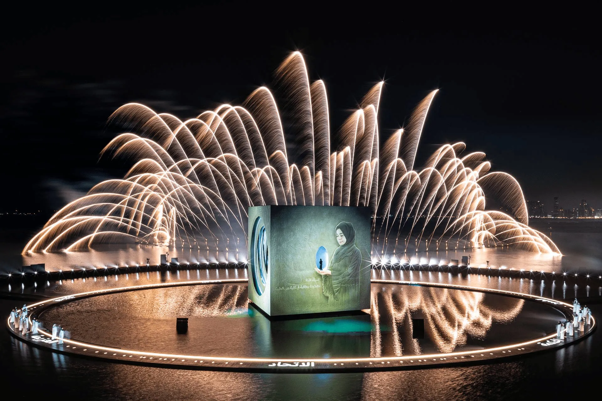 a giant cube standing on dark water at night, a woman's portrait projected on one face, fireworks fanning out behind it and performers in white lining the circular stage around it